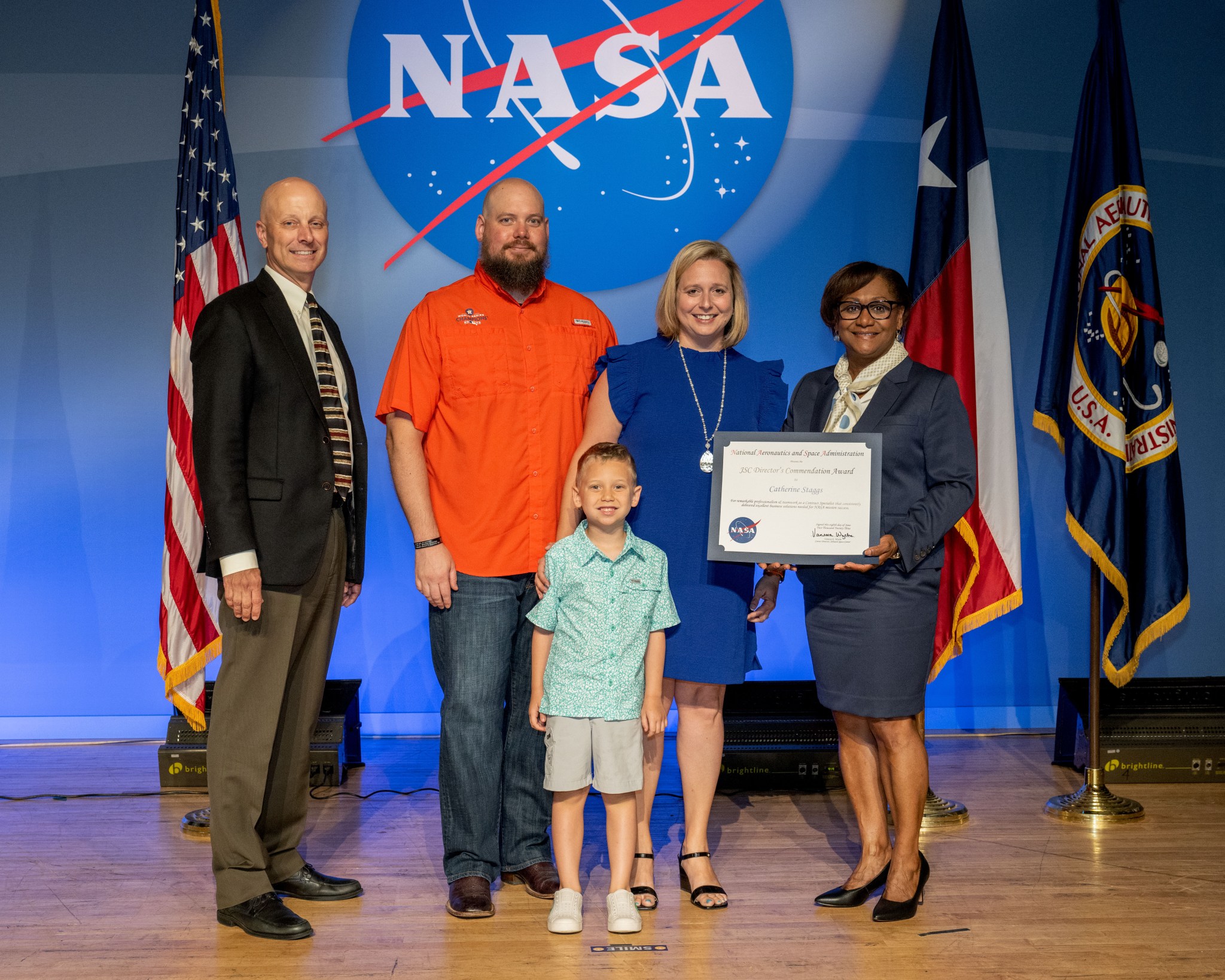 A blonde woman in a knee-length blue dress receives an award from the director of Johnson Space Center, on stage with her husband and young son.