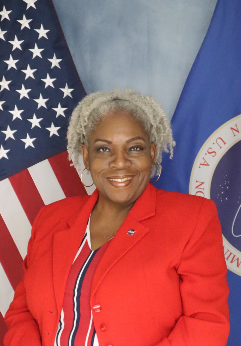 Image shows woman in red suit posing in front of American flag and NASA flag