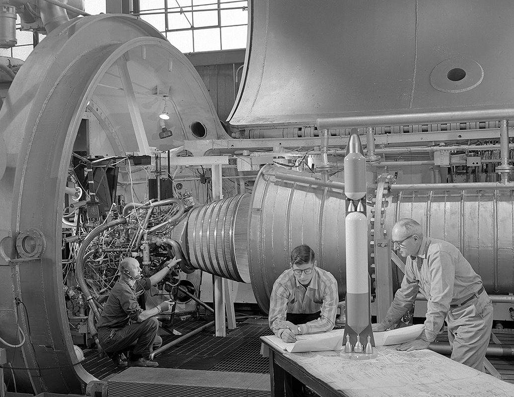 Men looking at rocket model with engine in the background.