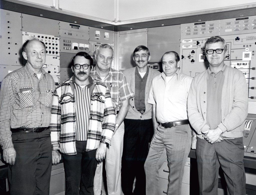 Group of men standing in control room.
