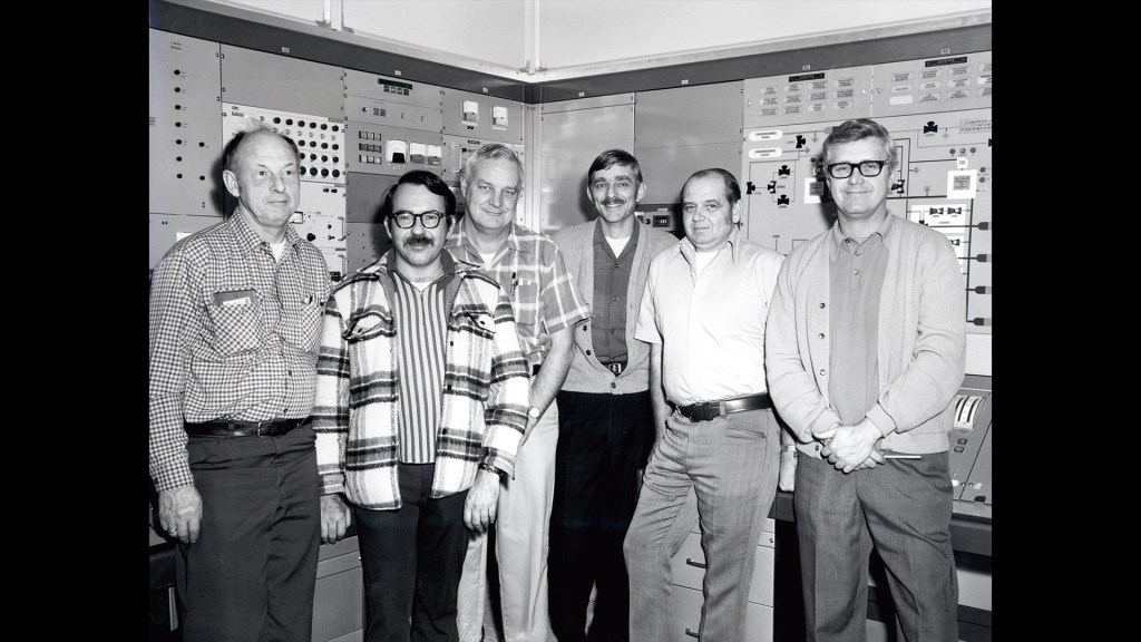 Group of men standing in control room.
