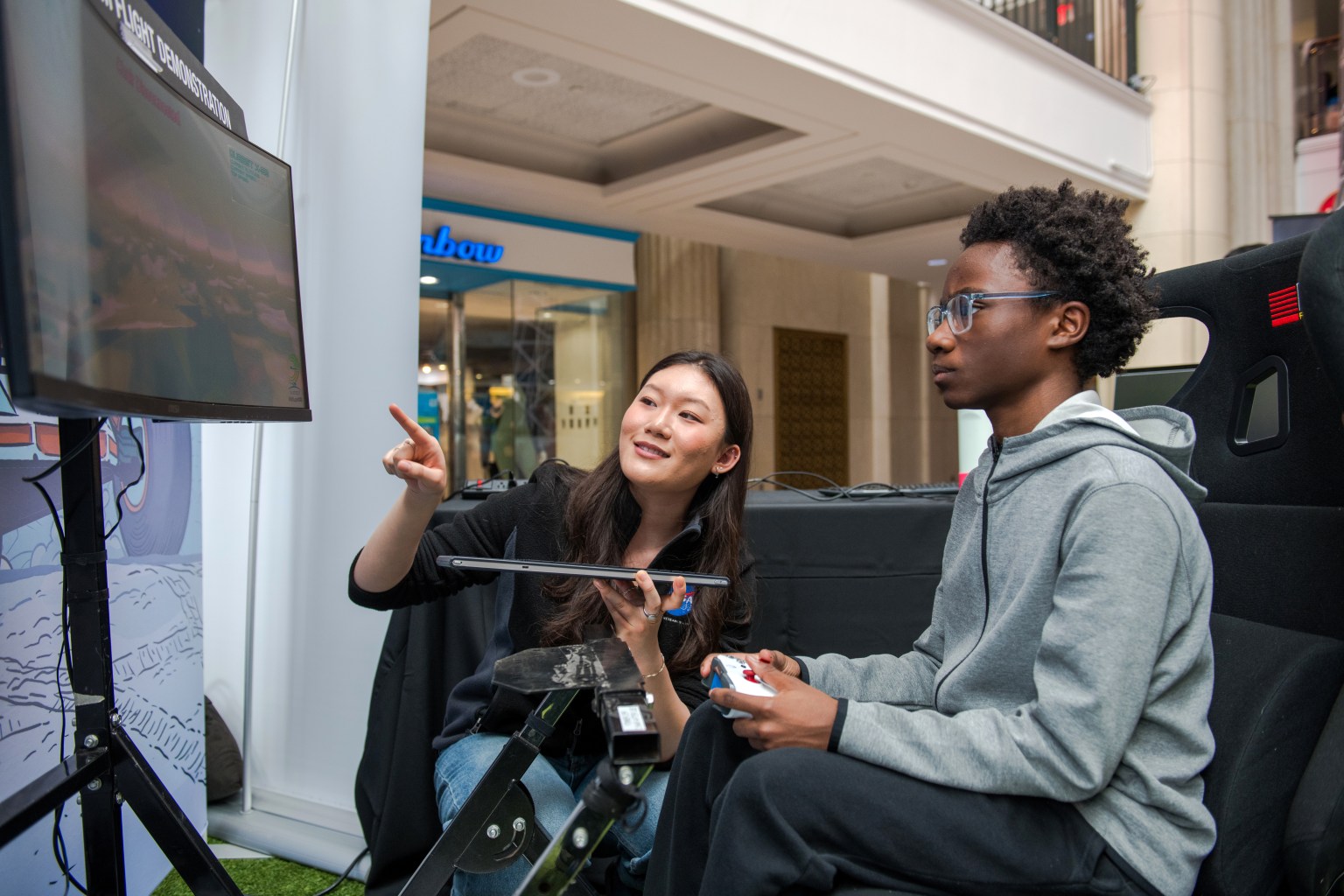A young boy with a controller in his hands looks intently at a television screen in front of him. A woman bends down next to him pointing at the screen, guiding him through the VR experience.