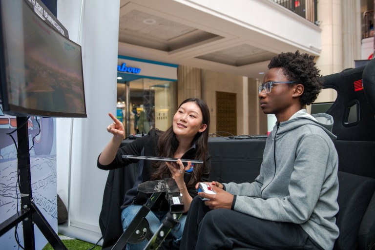 A young boy with a controller in his hands looks intently at a television screen in front of him. A woman bends down next to him pointing at the screen, guiding him through the VR experience.