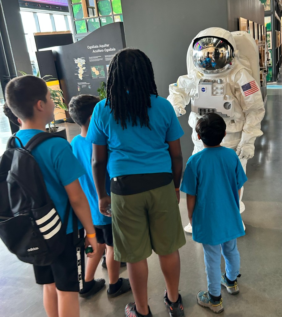 An astronaut mascot talks to a group of children wearing blue shirts. They are inside a building that highlights cultural information on Omaha, Nebraska.
