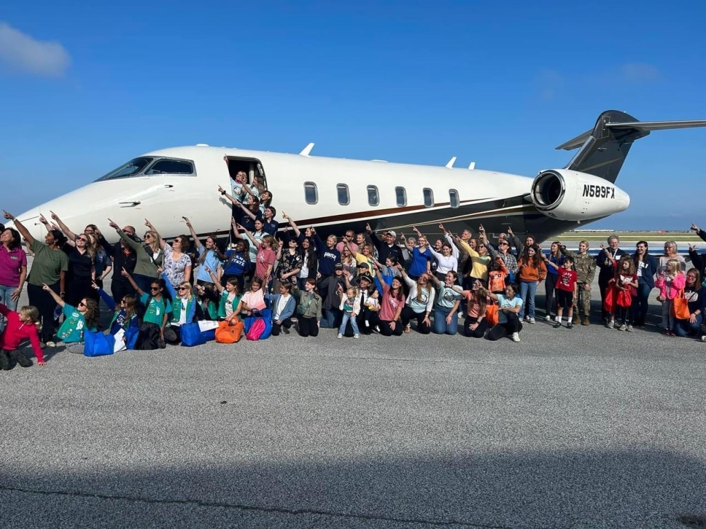 A large group of women and young girls gather in front of a large, white plane.  They stand outside and all point up to the left, signaling towards the sky.