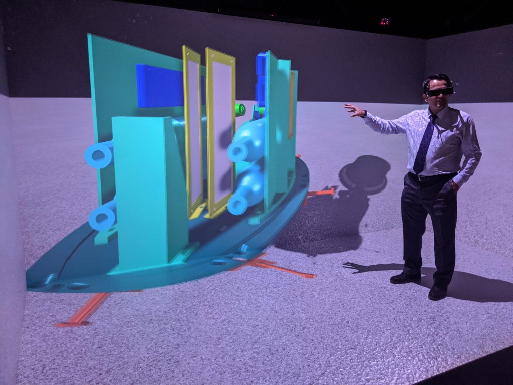 A man wearing a headset gestures towards a colorful 3D visualization.  He stands in a CAVE environment, so the walls around him and the floor beneath him all light up to display this VR experience.