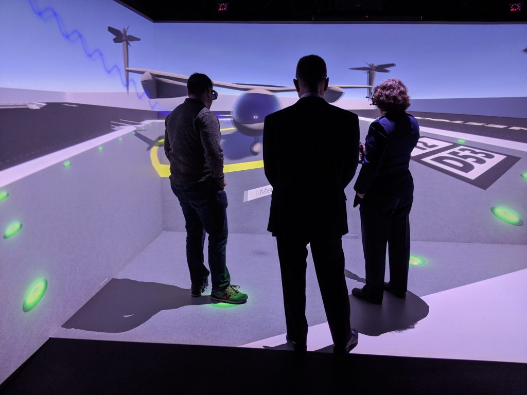 Two men and a woman all wearing headsets stand in a bright room viewing a NASA aeronautics visualization.  The walls around them and the floor beneath them all light up to display this VR experience.