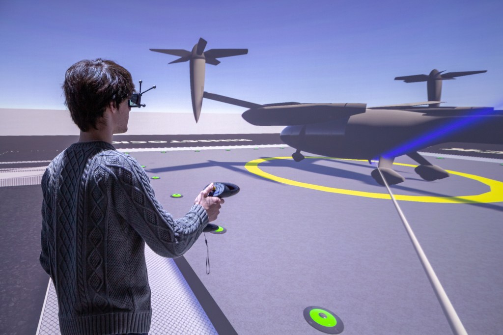 A man with a VR headset on his head and controller in his hand looks at a visualization of a plane sitting in a concrete lot.