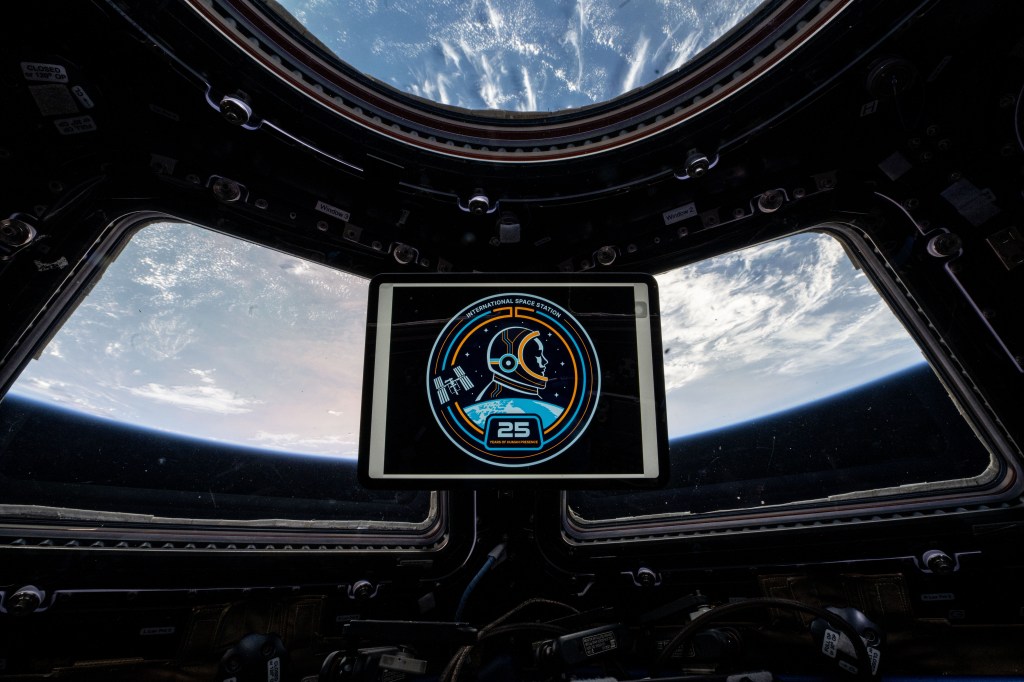A tablet displaying the logo created in celebration of 25 years of continuous human presence aboard the International Space Station floats in front of the cupola windows. Earth can be seen through the windows behind and above the tablet.