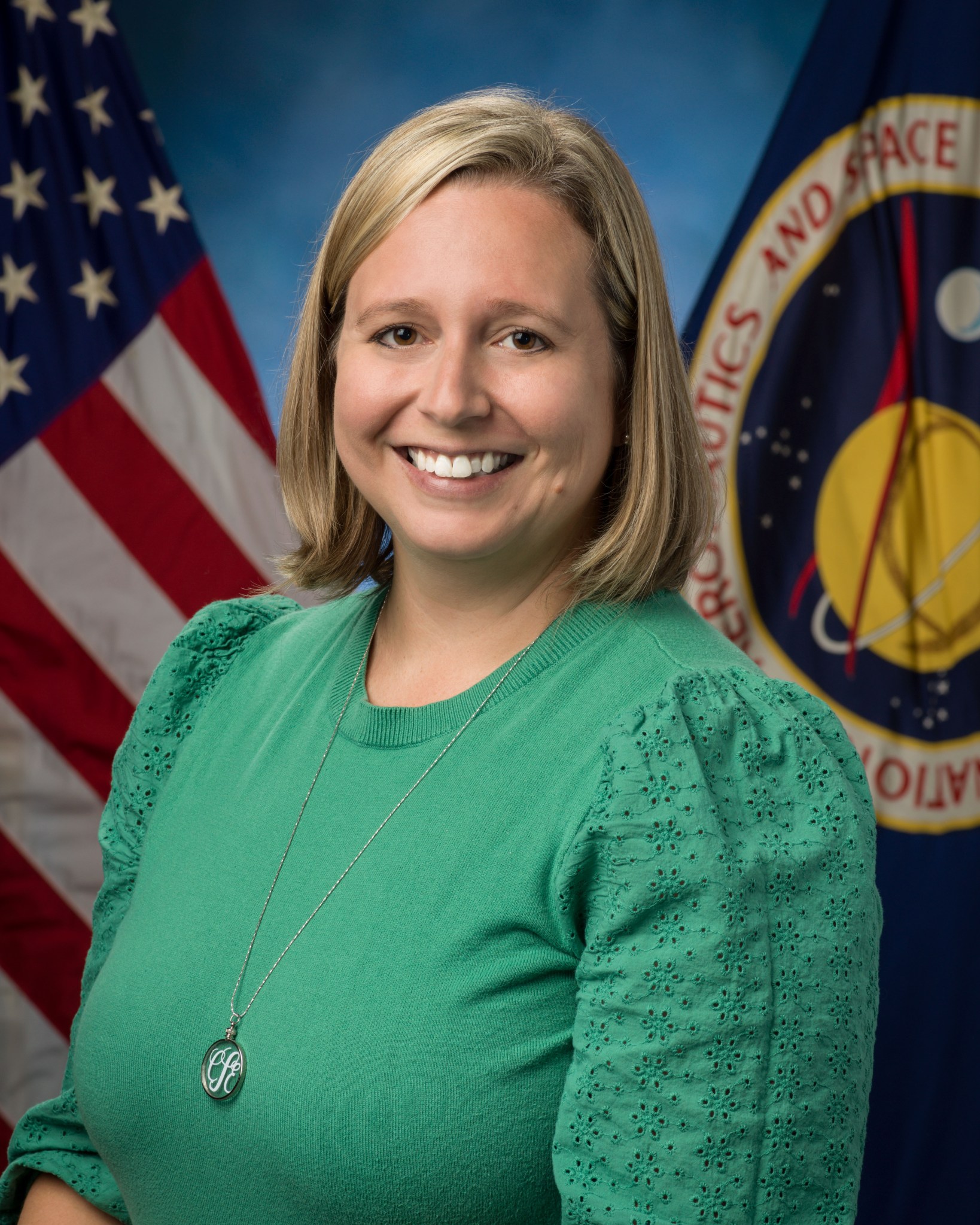 A woman with shoulder-length blonde hair, wearing a kelly-green blouse, stands in front of a NASA flag and an American flag in an official portrait.
