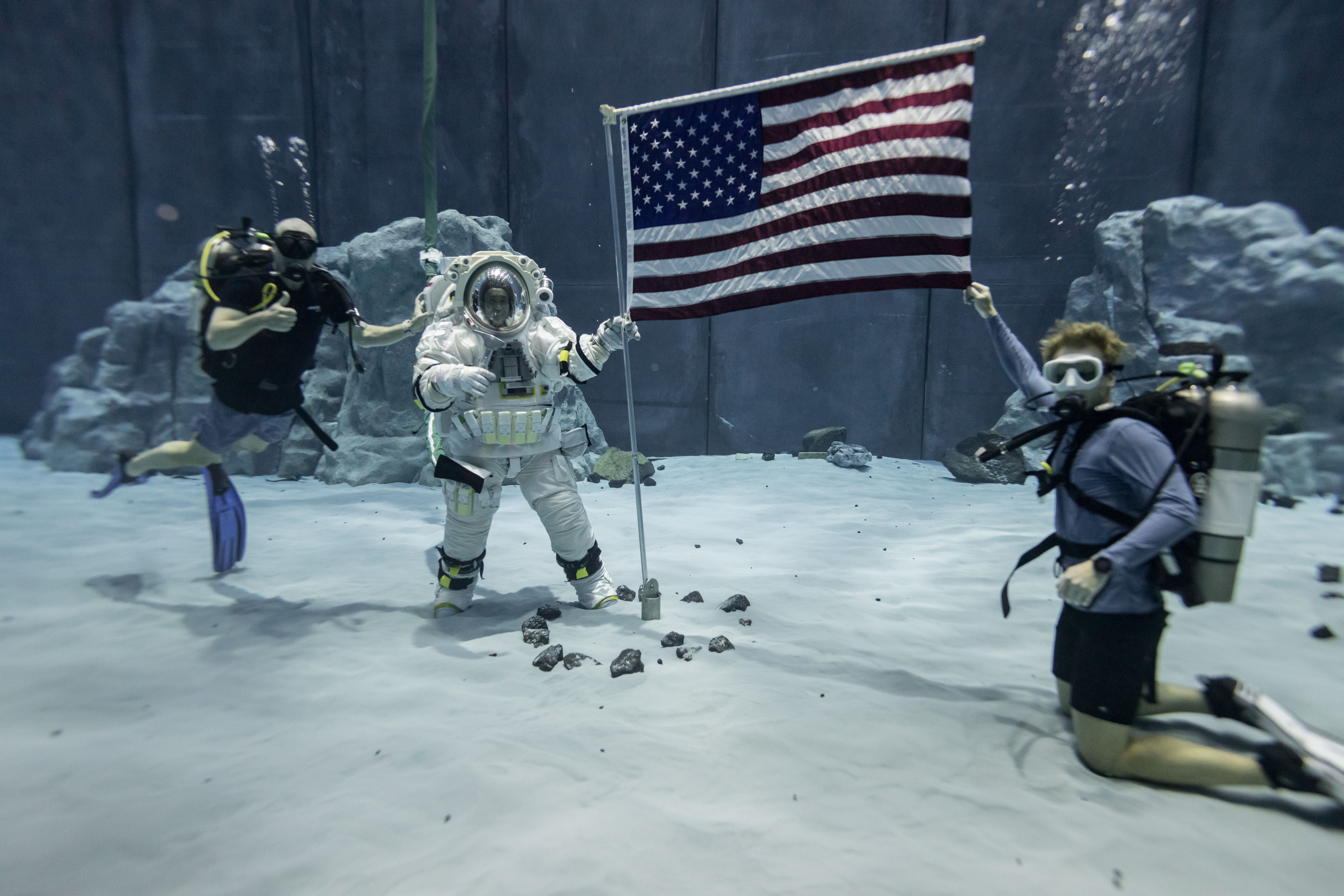 Axiom Space astronaut and Chief Technology Officer Koichi Wakata holds up an American flag while wearing a white spacesuit underwater with two scub...