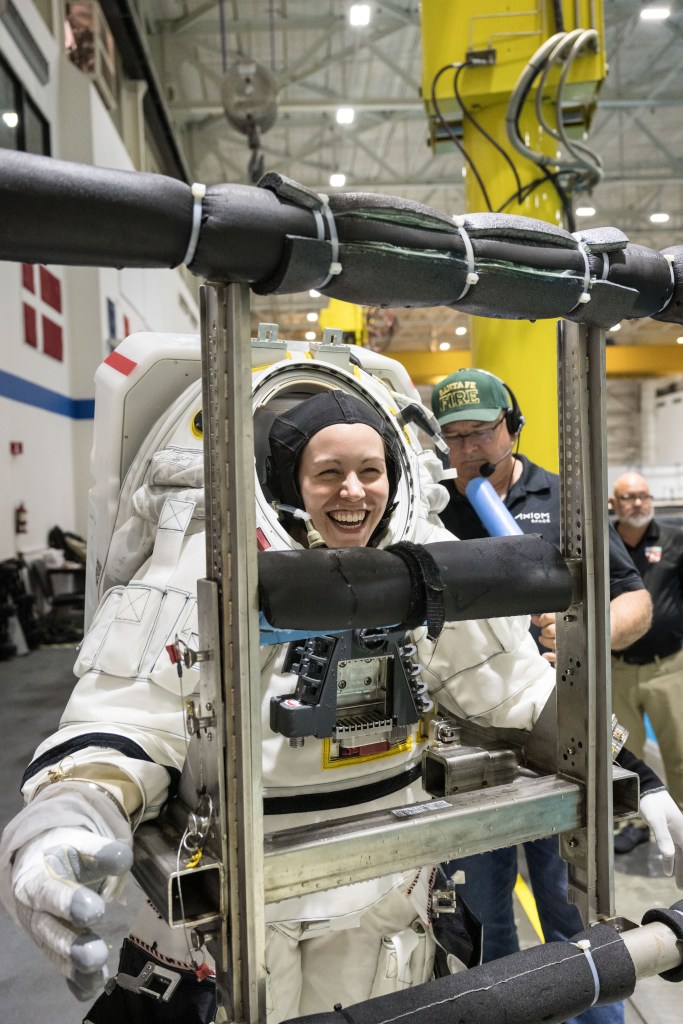 NASA spacesuit engineer Kristine Davis smiles big while wearing a white spacesuit that is on a metal donning stand that the spacesuit is attached to.