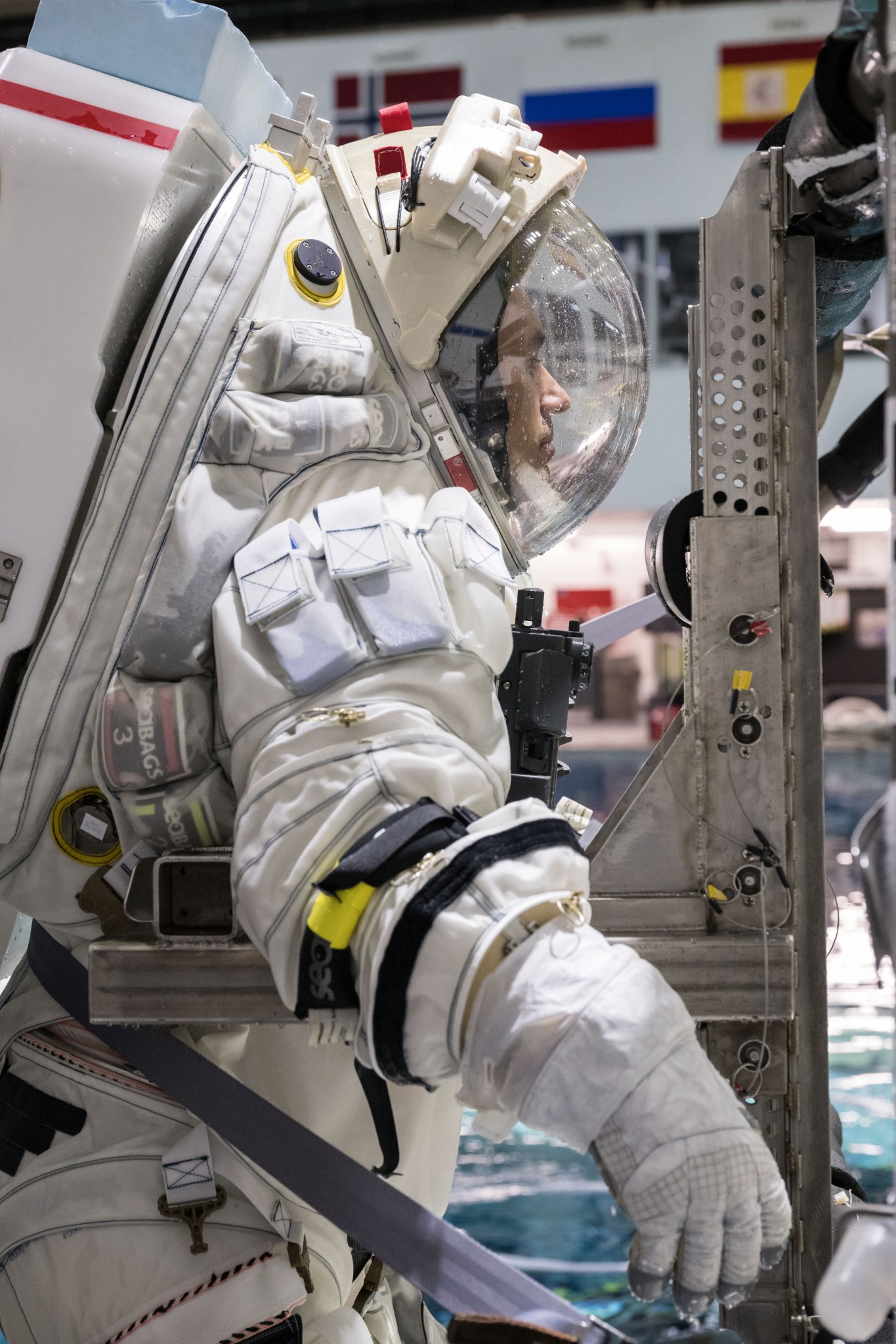 A side view of NASA spacesuit engineer Richard Rhodes wearing a white spacesuit that is attached to a metal donning stand. The helmet is closed and you can see water droplets on it.