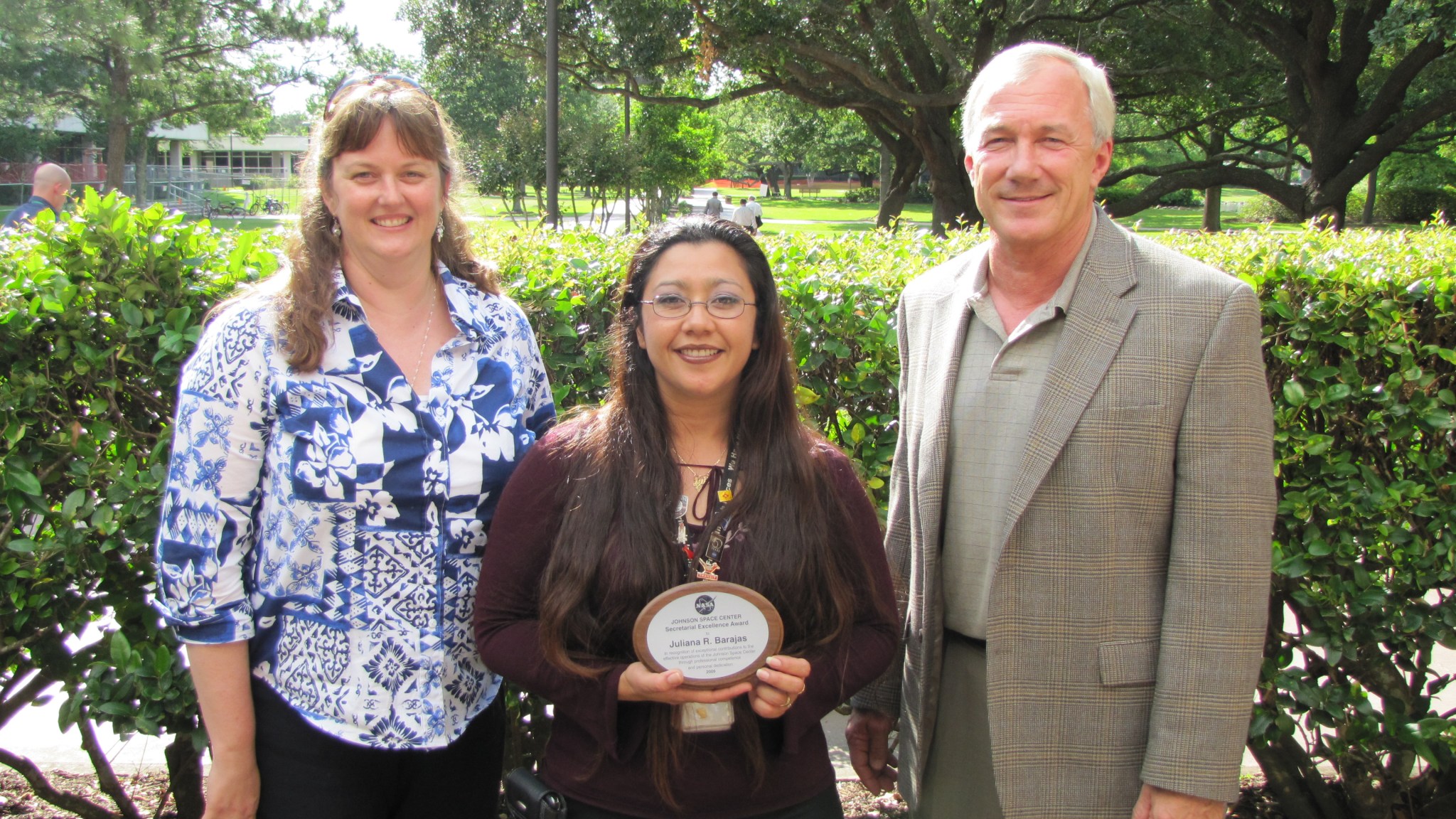 Three people stand outside in front of a bush and trees. The woman in the middle is holding a plaque.