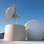 A white parabolic antenna atop a pedestal at NASA's Kennedy Space Center Uplink Station. A radome, or an enclosed dome housing another antenna, stands behind it. A blue sky and utility buildings are visible in the background.