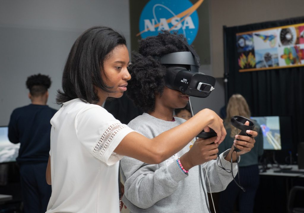 A young girl wearing a VR headset holding a controller experiences a virtual environment.  A woman standing next to her moves the controller, demonstrating how to use the device.