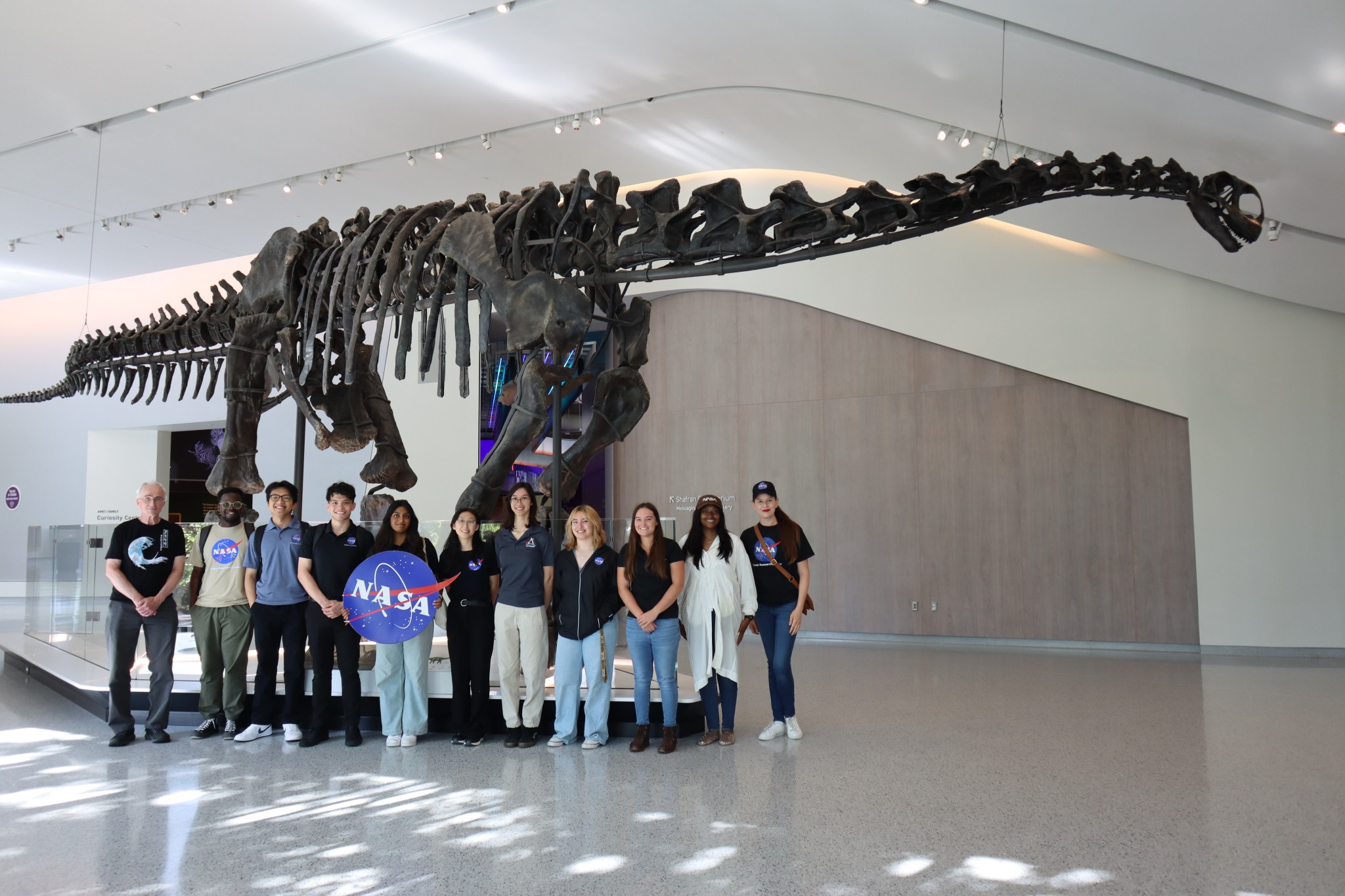 A large group of NASA employees stand smiling in front of a skeleton of a dinosaur.  One employee in the middle holds a sign reading: "NASA".