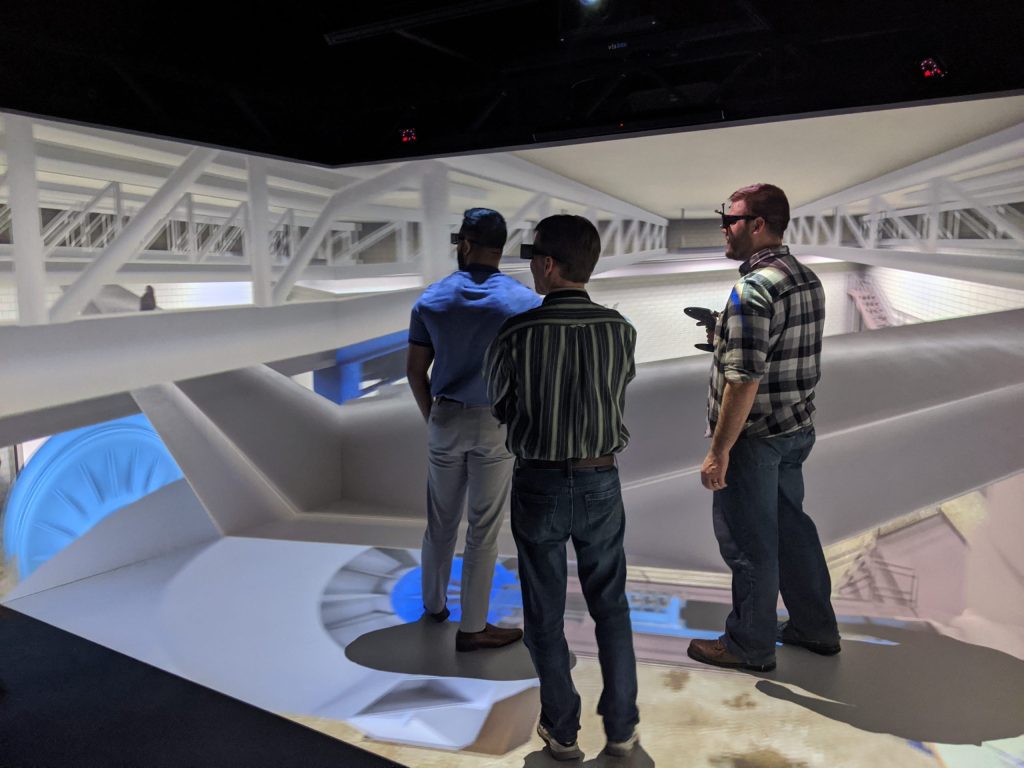 Three men wearing headsets and holding controllers stand in a bright room that shows a 3D visualization.  The walls around them and the floor beneath them are screens and display a VR version of a real NASA facility.