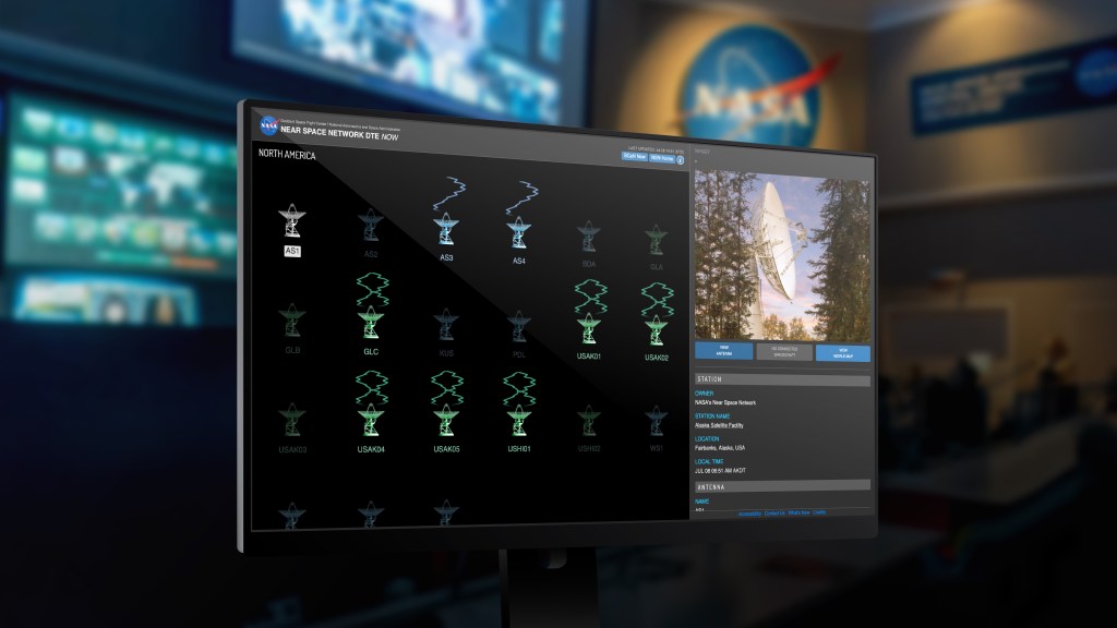 Computer monitor displaying NASA’s Near Space Network DTE NOW interface with icons of ground antennas across North America. The screen highlights the Alaska Satellite Facility (AS1) with a photo of its dish antenna and location details in Fairbanks, Alaska. NASA and Near Space Network ground station maps appear blurred in the background control room.