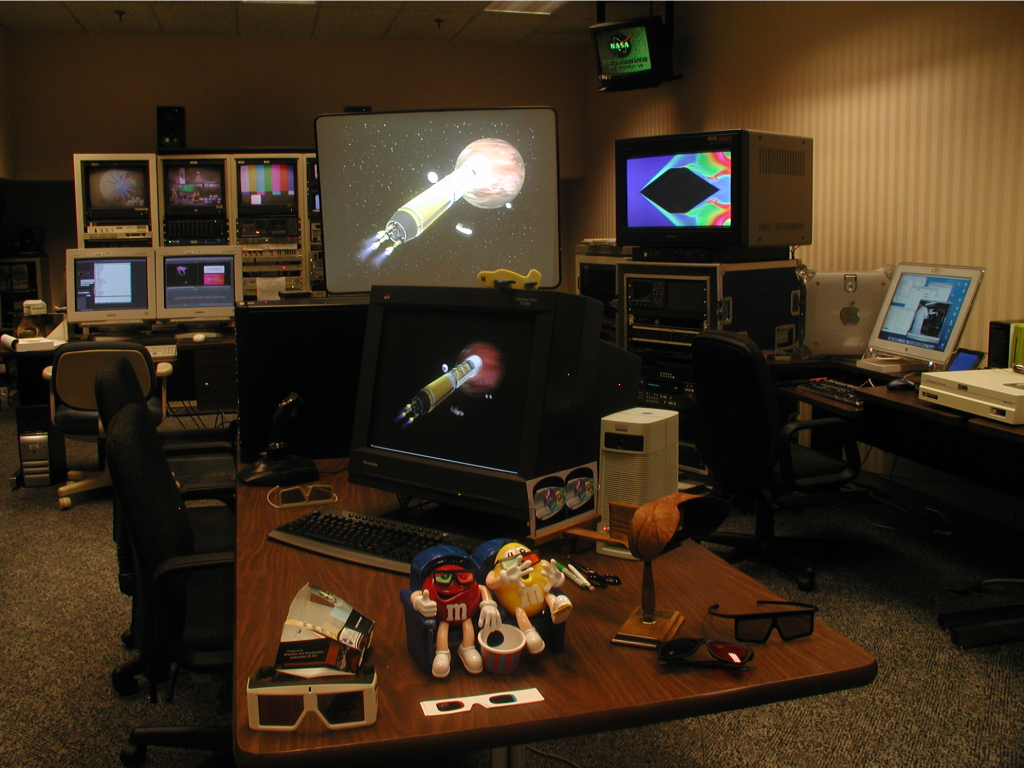 A table with VR glasses, a computer, and other equipment sits in the center of a darkly lit, cramped room.  Various computer and television screens line the walls behind the table.