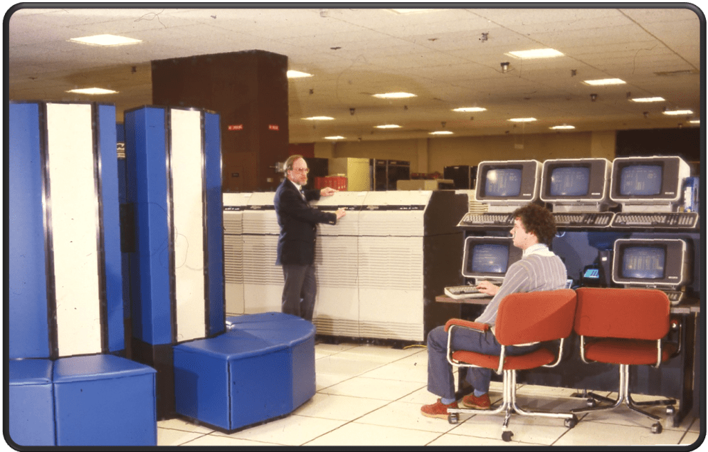 Two men face each other talking in a bright, windowless office room.  Computer screens stacked on top of each other sit to the right and tall equipment sit to the left.