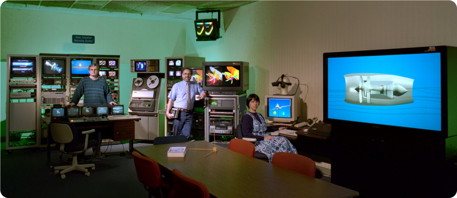 Three NASA employees sit in a large room filled with glowing television and computer screens.