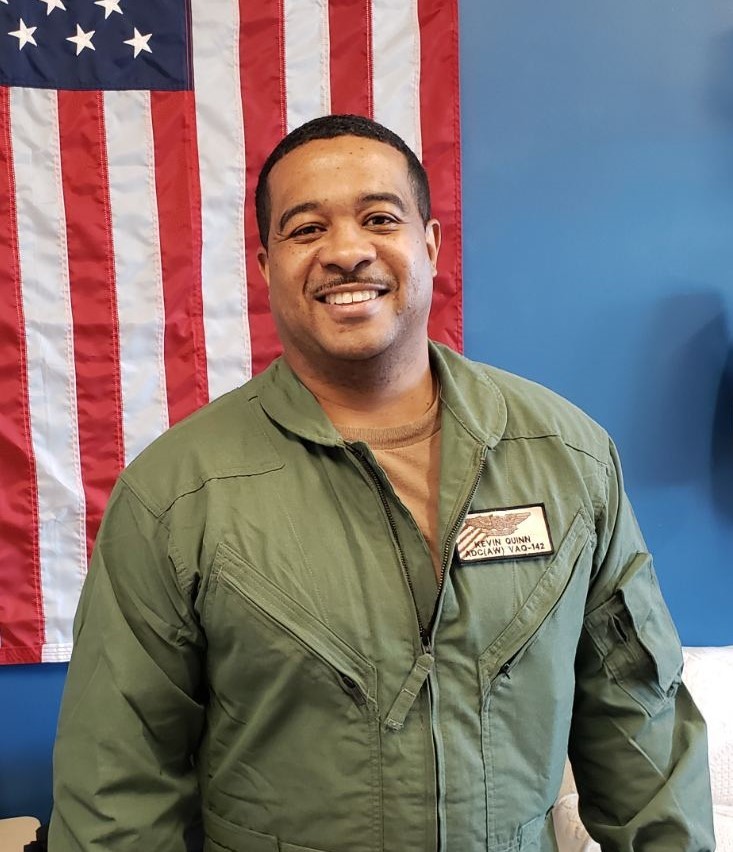 Black man wearing a military-green flight suit stands in front of an American flag.