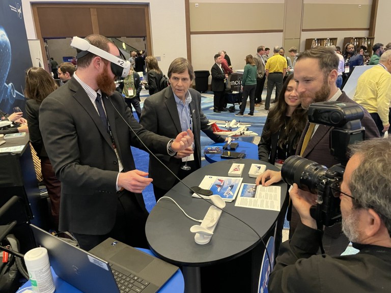Group of people around a table. One has a virtual reality headset on while the rest observe.