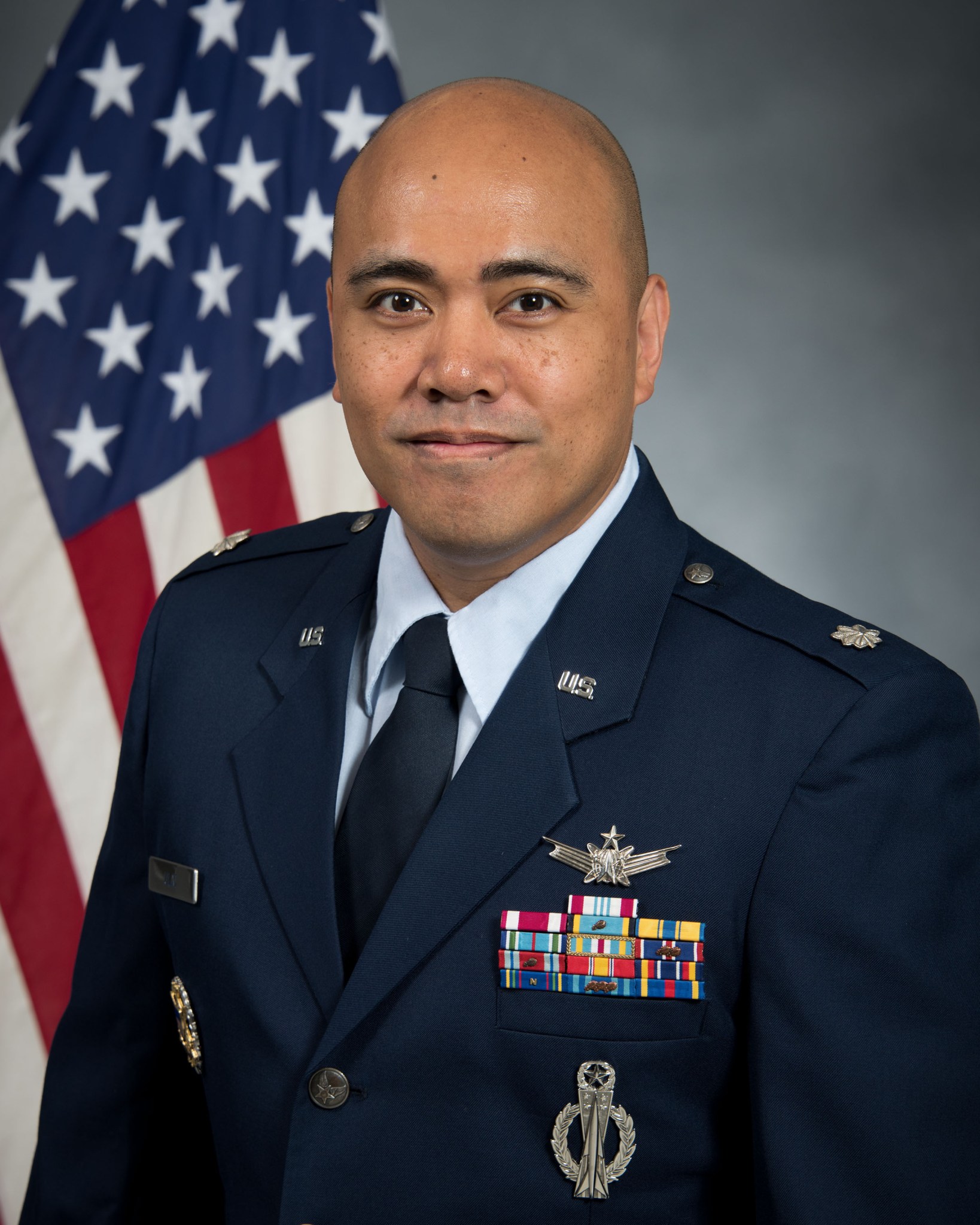 A bald Asian man wearing an Air Force dress uniform stands in front of an American flag.