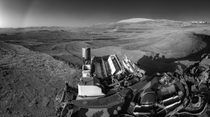 A black and white photo of the Curiosity rover on Mars. Curiosity is at bottom right. The rover's nuclear power source is visible; it is lined with rows of white fins at the back of the rover. Tracks trail off into the distance. Curiosity's landing site is on the horizon at top left. Mars' terrain is rocky.