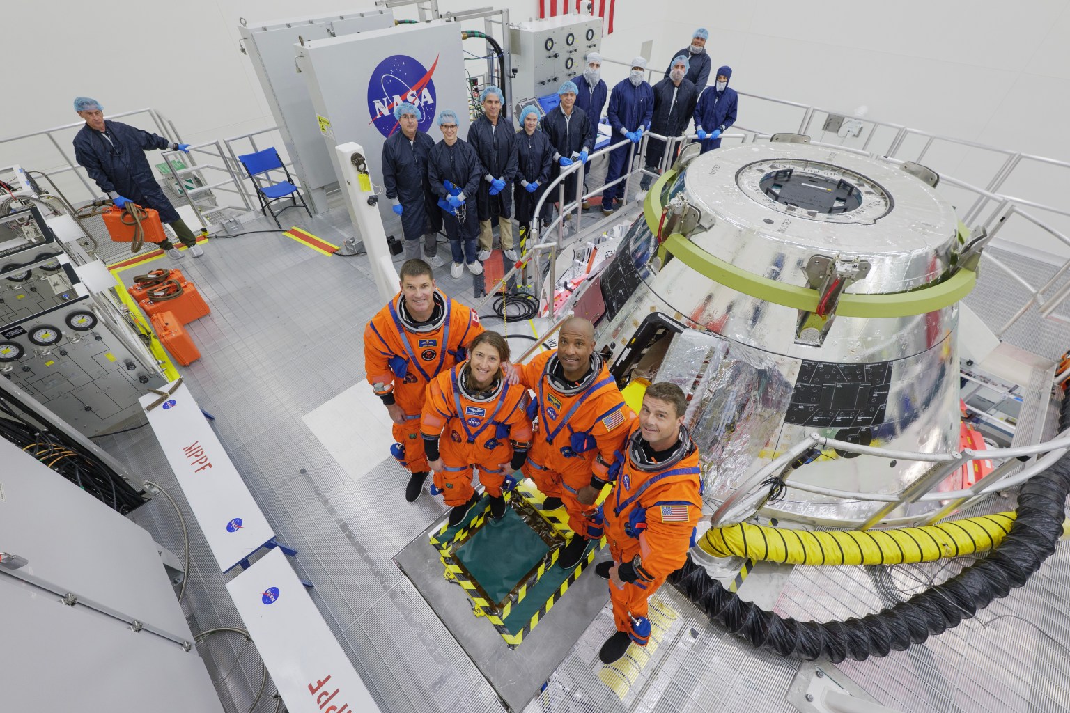 The Artemis II crew stands in front of the Orion spacecraft. They are wearing orange spacesuits without helmets. Orion is silver-colored and reflective. There is a round railing surrounding the spacecraft. There is also a group of people wearing blue jumpsuits, hairnets, and gloves standing behind Orion; they are members of the Artemis II closeout crew.