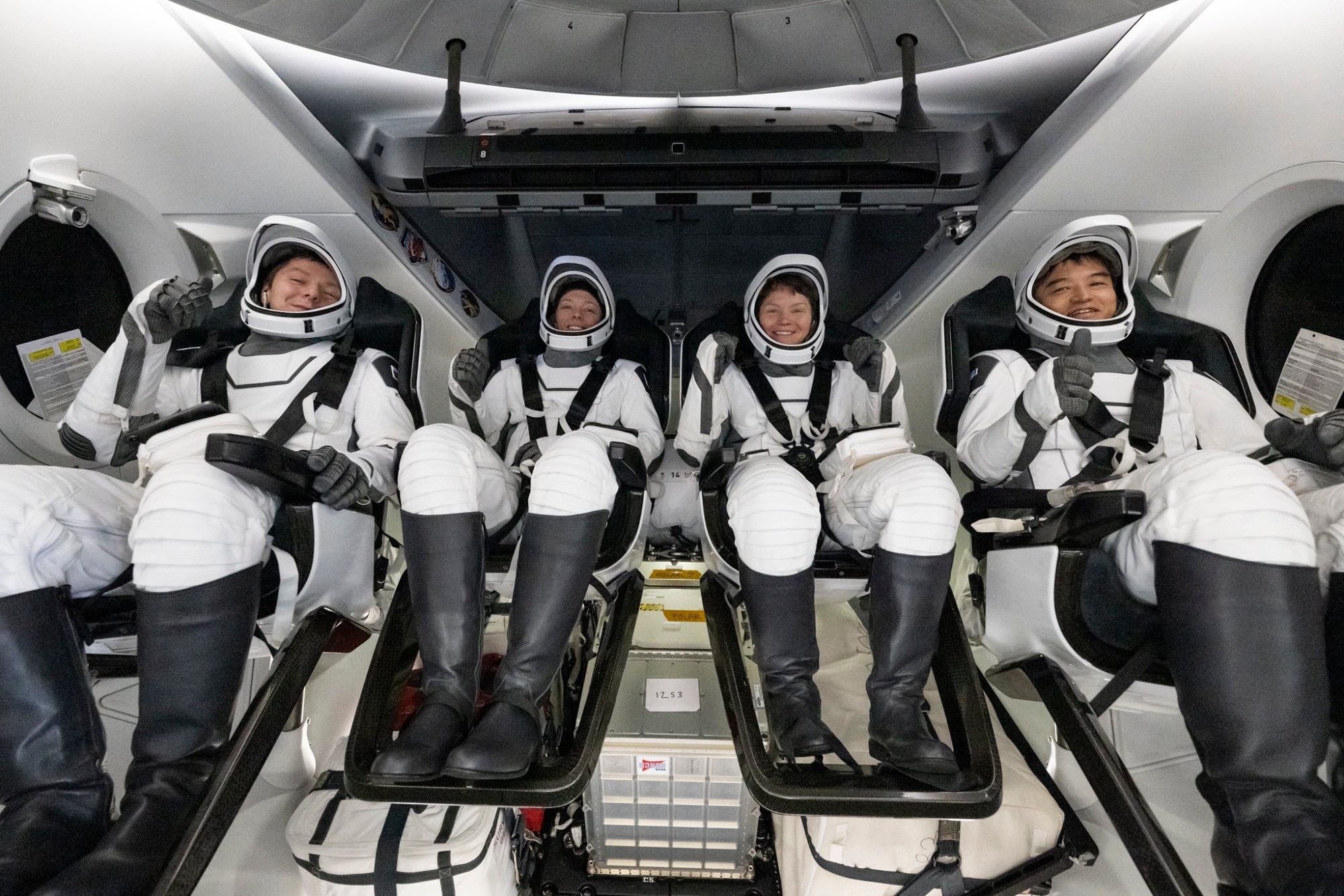 Four astronauts in white flight suits and black boots are smiling, seated inside a white and black spacecraft.