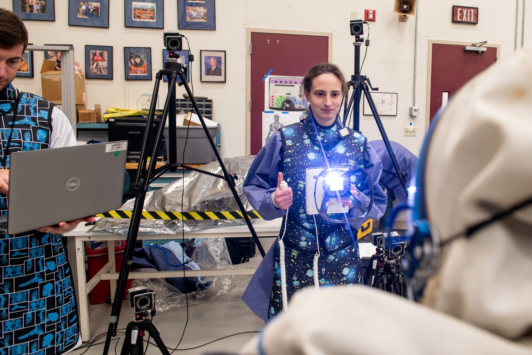 A researcher in a lead apron clicks the button on a portable handheld X-ray system to capture an image of a spacesuit. A flash of light beams from the system. A researcher stands next to her in the lab peering down at his computer. Behind them are two cameras on tall tripods and one small camera near the ground. On the walls are photos of NASA astronauts and posters displaying astronaut health research imagery.