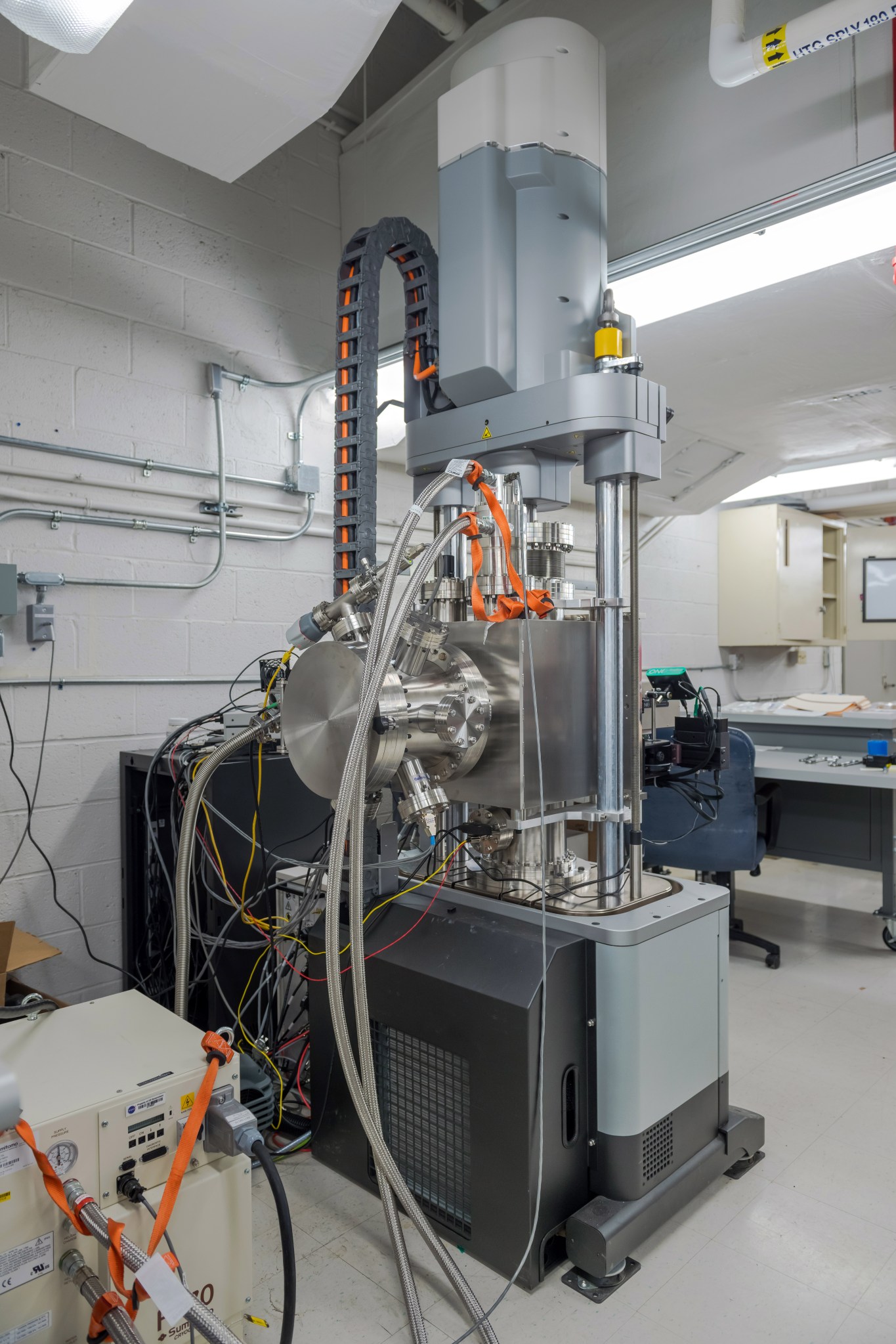 A tall, industrial machine with metallic and gray components stands in a laboratory. Multiple cables and tubes connect to the central chamber, which is cylindrical. Some cables are silver and braided, while others are orange or yellow. The test rig has vertical support columns and a black base. In the background there are white brick walls, ceiling ducts, and a work station with a desk and chair.
