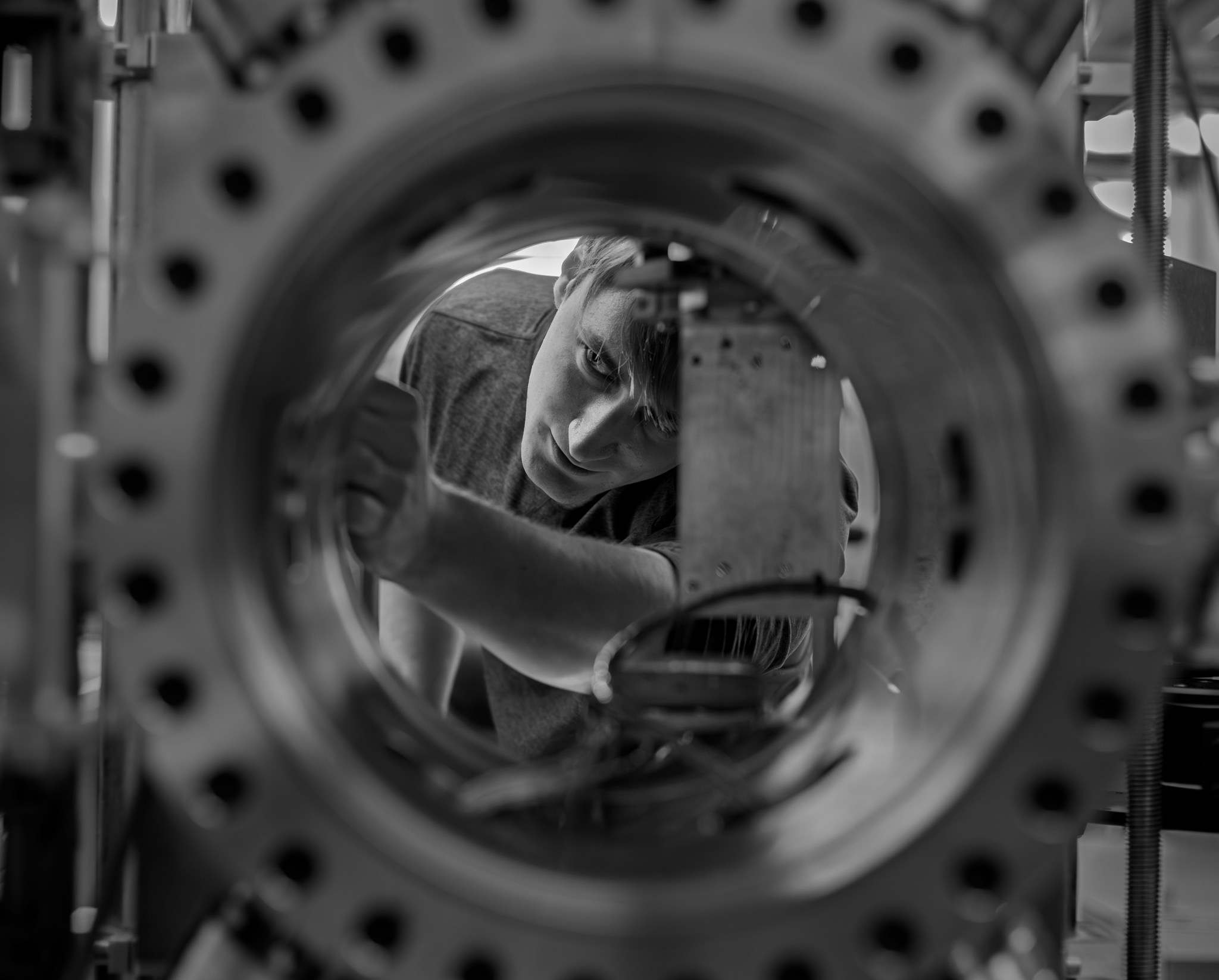 A black-and-white photo of a cryogenic engineer viewed through a circular opening of a large metallic chamber with evenly spaced bolts along its rim. Inside, a square component is mounted with wires connected to it. The man viewed through the porthole appears to be adjusting settings for the machine, which can mimic the extreme cold of the lunar night. 