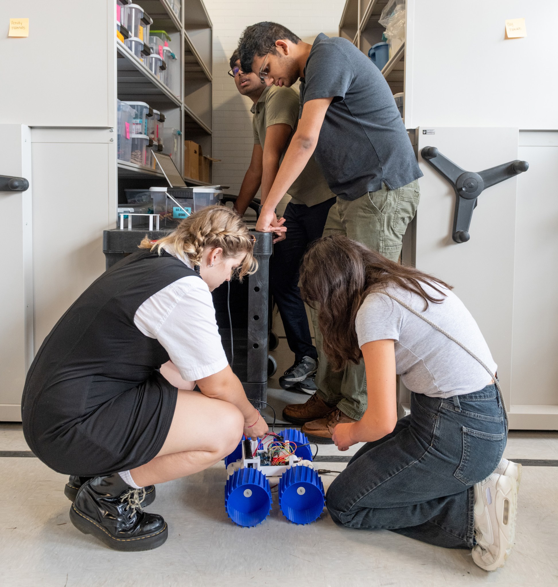 Two students kneel and adjust wires in a small rover with large blue wheels while two other students stand between cabinets containing several clear plastic containers