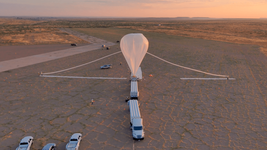 Aerial view of a high-altitude scientific balloon launch preparation in a desert setting at sunset. The white balloon is partially inflated and tethered to a launch vehicle, with support vehicles and personnel positioned around the launch area on a cracked desert surface