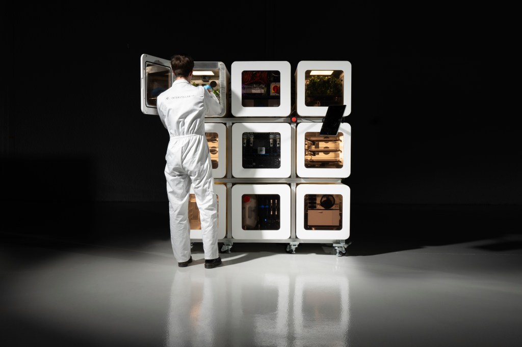 A person in a white jumpsuit stands in front of Interstellar Lab's NuCLEUS technology, comprised of nine food-producing chambers arranged in a 3x3 stack.