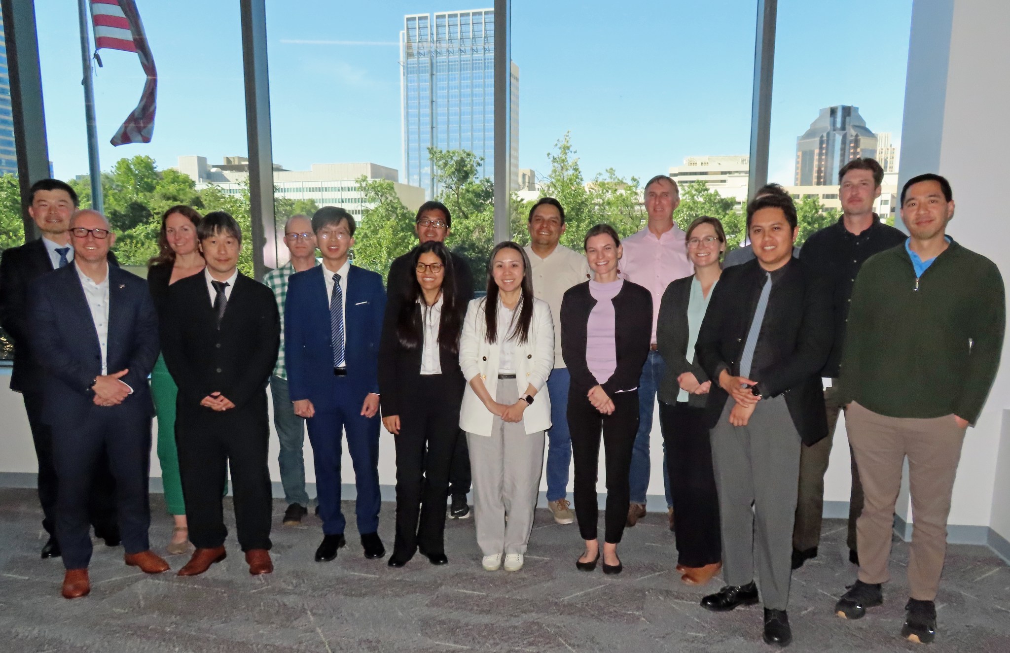 A group of about 15 people post together in front of large windows, with blue skies and a large skyscraper visible in the distance.