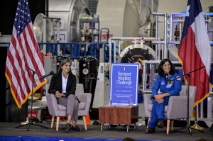 Second Lady Usha Vance and astronaut Suni Williams sit in gray chairs on a stage. They are both smiling as they listen. The U.S. flag, the state flag of Texas, and a flag with the NASA seal are on the podium alongside them. There is a blue poster board with a white border promoting the Summer Reading Challenge between them.