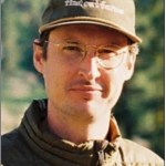 Headshot of a man wearing clear glasses, a brown baseball cap and brown puffy coat.