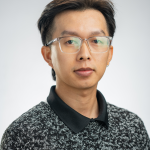 Headshot of a young man with short black hair, clear-rimmed glasses, and a black speckled shirt.