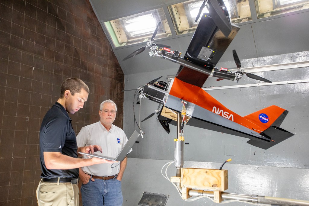An orange-colored airplane research aircraft sits in a wind tunnel facility mounted on a strut. The aircraft consists of a section of a scaled-down fuselage with two wings and six rotors. Two researchers are using a computer connected to the aircraft. The gray wall of the wind tunnel is visible in the background.