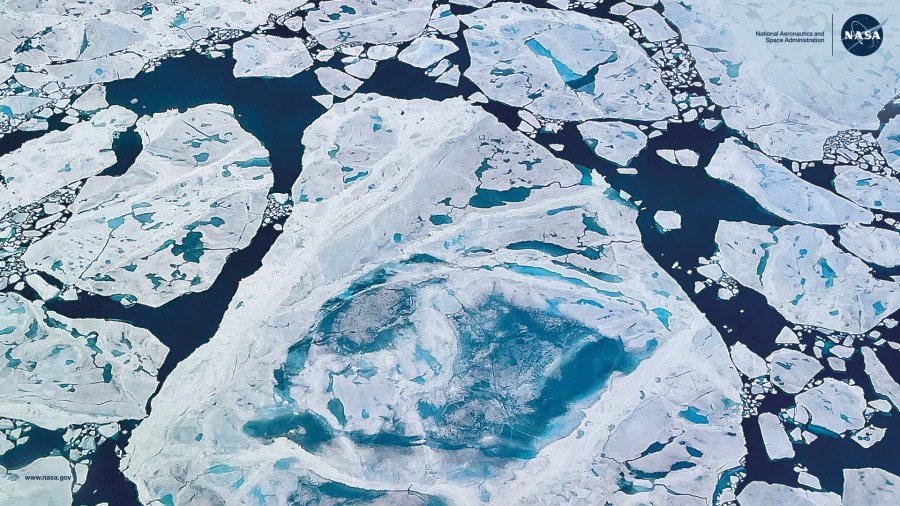 Aerial view of fractured sea ice floating in the Arctic Ocean. Large white ice sheets are broken into irregular shapes, with deep blue melt ponds and dark open water channels visible between them.