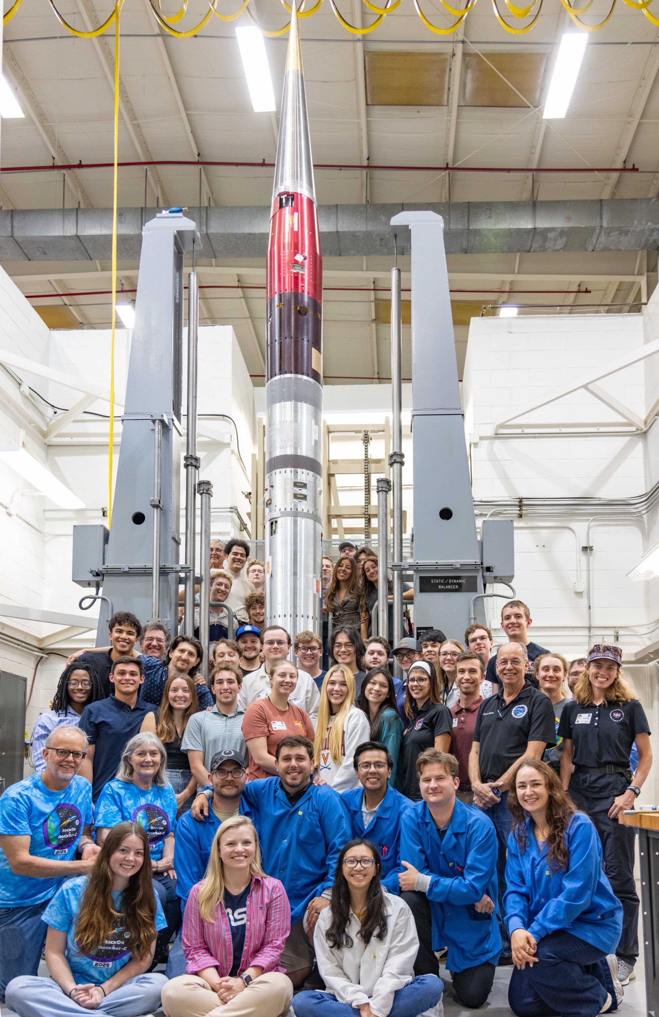 "A group of people gathered in front of a sounding rocket in an assembly facility. The silver and red rocket stands vertically between support structures in a bright indoor space with white walls and yellow ceiling cables. University student team members wearing blue lab coats are positioned in group photo formation in front of the rocket."