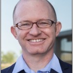Headshot of a bald man wearing glasses, with a blue collared shirt and dark blazer.