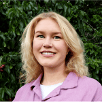 Headshot of a young white woman with medium-length blonde hair, wearing a lavender buttonup shirt and standing against dark green foliage.