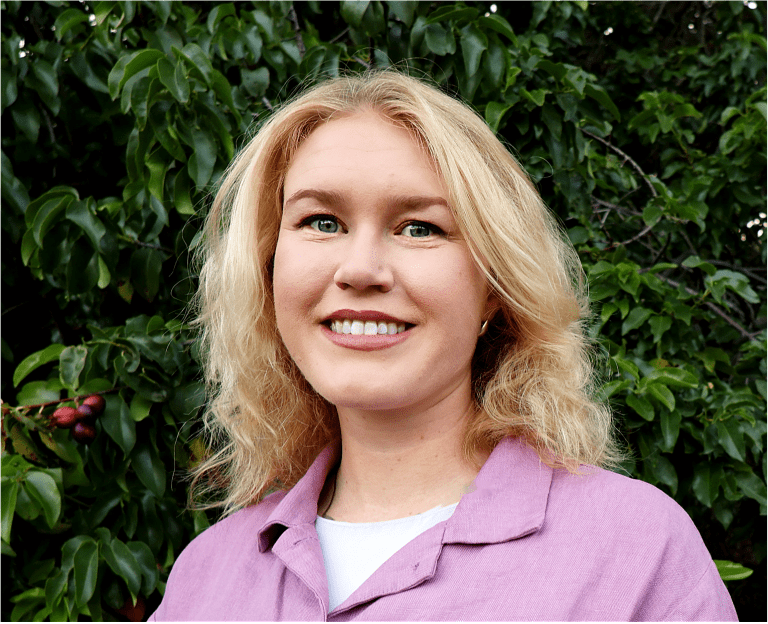 Headshot of a young white woman with medium-length blonde hair, wearing a lavender buttonup shirt and standing against dark green foliage.
