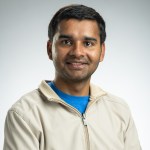 Headshot of an Indian man with short black hair, wearing a cream colored jacked and bright blue shirt.