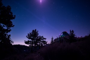 A faint beam stretches up and to the left from the open dome of a laboratory into the sky. The building and surrounding trees are mostly in shadow. The sky is a deep blue with multicolored stars visible; the bottom of the sky where it meets the landscape fades from blue to pink.